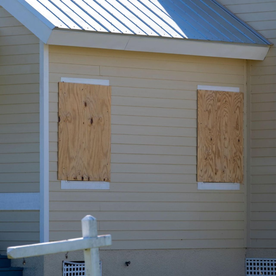 Beige house exterior with two boarded-up windows and a white fence in front