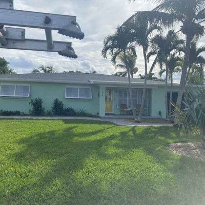 A single-story light green home with a dark roof and a wide green grass lawn, featuring white-framed windows and palm trees in the background.