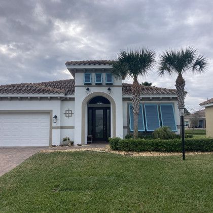 A Mediterranean-style luxury home with a white garage door, an arched front entrance, and light blue shutters on the front windows, flanked by two palm trees.