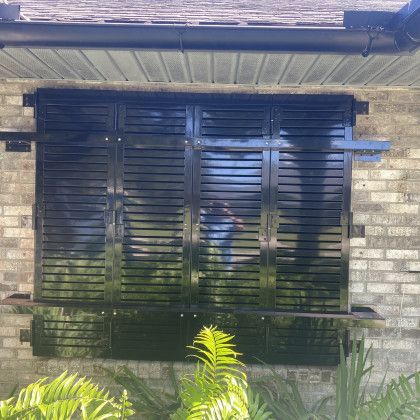 A close-up shot of a window on a brick house covered by dark, glossy black louvered shutters reflecting the outdoor light.