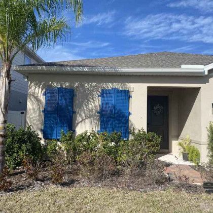 Close-up of a tan stucco house featuring bright royal blue louvered shutters closed over two front windows, surrounded by landscaping.