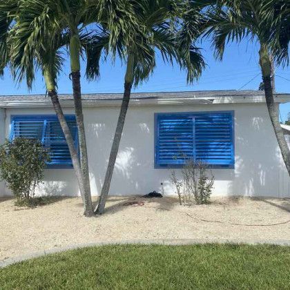 The front facade of a white house partially shaded by palm trees, featuring two windows with vibrant blue Bahama-style storm shutters.