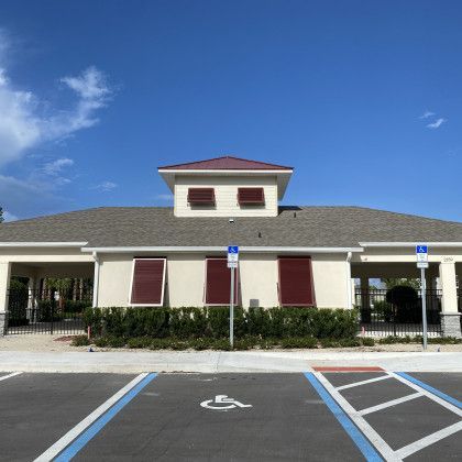 A large commercial or community building with a grey shingled roof, featuring deep red or burgundy hurricane shutters on all windows, viewed from a parking lot with a handicap space.