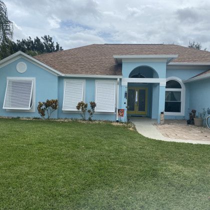 A light blue single-story house with a green lawn, featuring white louvered storm shutters on the windows and a prominent central arched entryway.