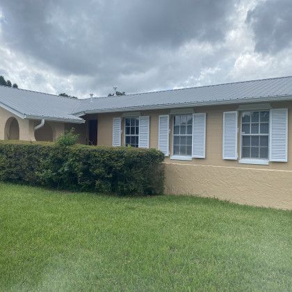 An angled shot of a tan house with a metal roof and a row of green bushes, featuring white decorative shutters on multiple windows.