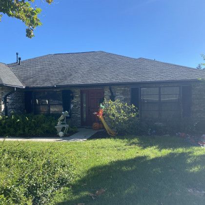 A single-story home with dark siding or stone accents and a dark roof, surrounded by green lawns and trees under a bright blue sky.