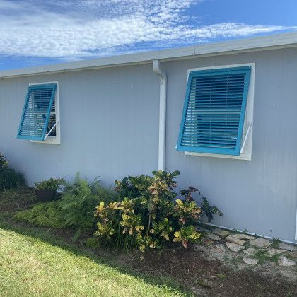 Side profile of a light blue or grey house featuring two turquoise-colored Bahama shutters propped open over the windows.