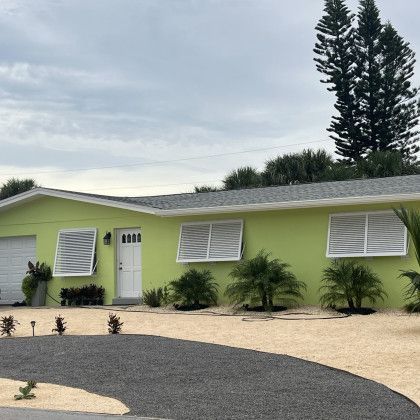 A lime green single-story house with white trim, a dark curved driveway, and white horizontal shutters over the windows.