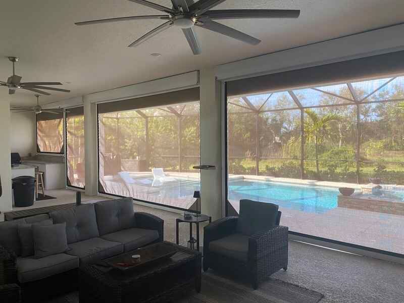 View from inside a covered patio looking out toward a swimming pool, featuring wicker outdoor seating, a ceiling fan, and large motorized screens lowering over the openings.