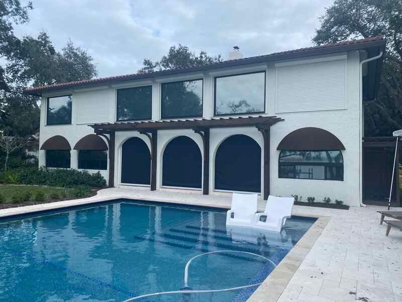 A luxury white two-story home with a swimming pool, featuring a pool deck with two white lounge chairs and dark fabric roll-down screens installed within arched patio openings.