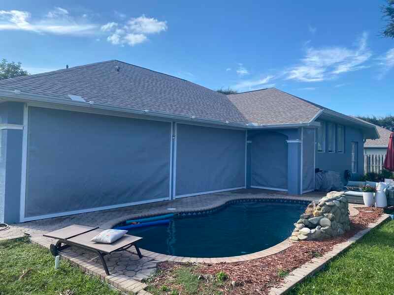 The backyard pool area of a single-story grey house, featuring large motorized privacy and sun shades completely rolled down to enclose the back patio.