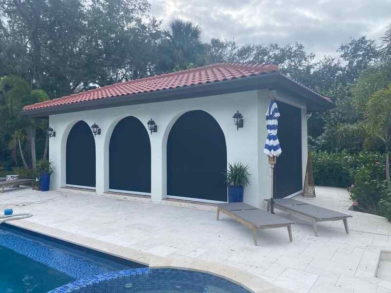 An outdoor pool cabana structure with a terracotta tile roof, white stucco walls, and black roll-down privacy screens fully lowered into the arched walkways.