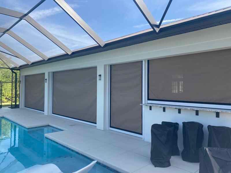 A white modern pool house or patio enclosure under a screen cage, featuring beige pull-down solar shades covering the glass doors and windows next to the swimming pool.