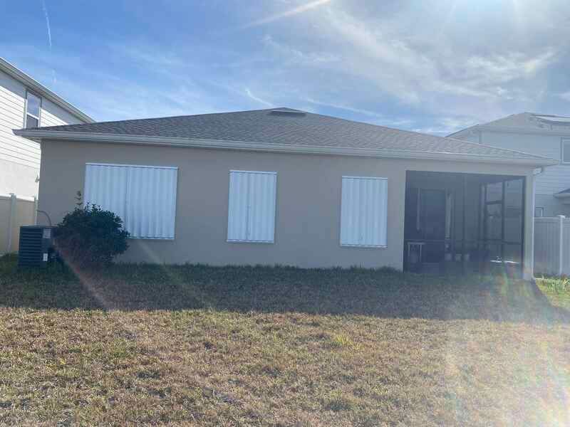 The same single-story tan home from Image 1, but with white accordion hurricane shutters completely closed over all three windows.