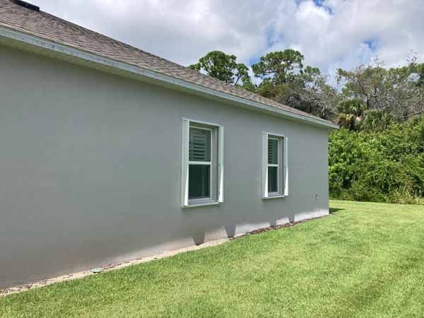 An angled side-profile shot of a light grey stucco house showing two white-trimmed windows and a green grass lawn.