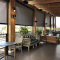 Covered wooden patio with wicker chairs, potted plants, and outdoor tables beside large roll-down shades