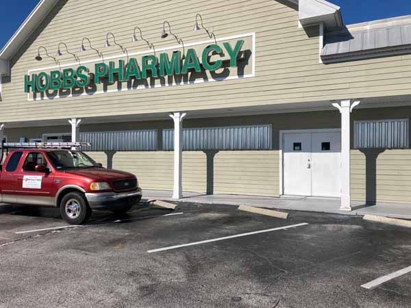 The front exterior of a commercial building with tan siding and a sign reading 'BBS PHARMACY', featuring clear corrugated plastic panels covering the long row of front windows next to a white double door, with a red pickup truck parked out front.