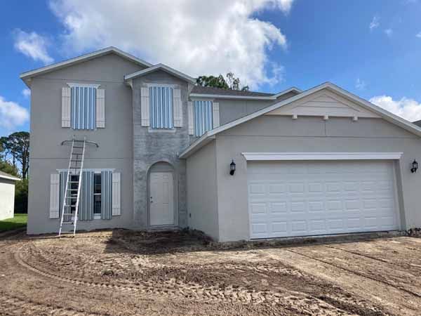 A two-story grey stucco house under construction with a dirt yard, featuring clear corrugated plastic storm panels installed over all the windows on both floors.