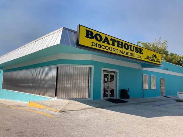 The exterior of a teal commercial building with a bright yellow sign reading 'BOATHOUSE DISCOUNT MARINE, LLC', featuring vertical silver metal hurricane panels covering the large storefront windows.