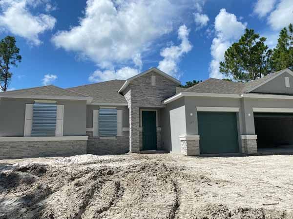 A newly constructed or renovated single-story grey home with stone accents, a dark green garage door, and clear corrugated hurricane panels mounted over the front windows.