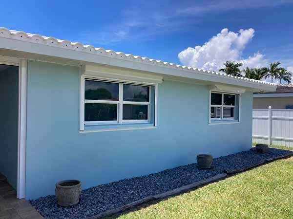 Blue house with two windows and retracted white hurricane shutters above them.