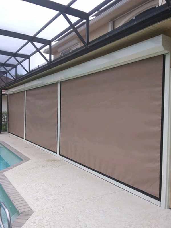 Covered patio with large tan motorized screens lowered next to a pool.