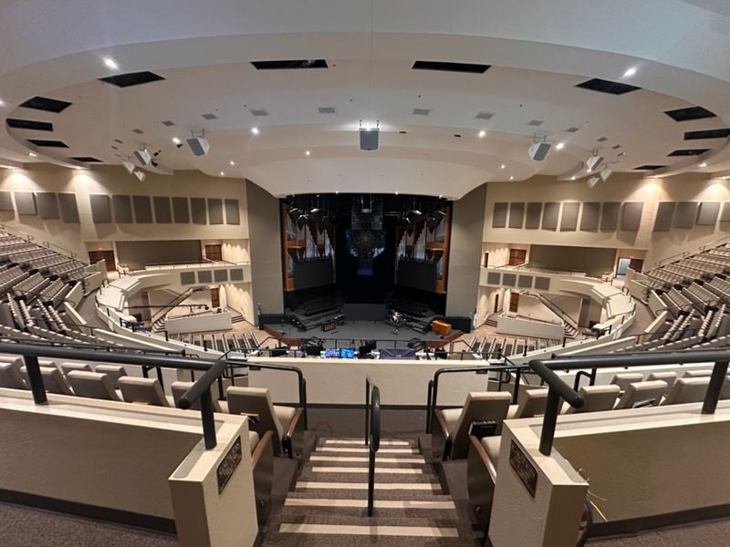 A high-angle, wide view of a modern auditorium featuring tiered, light-colored seating facing a central stage area.