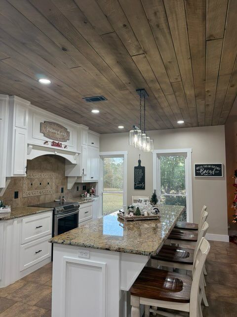 A kitchen with white cabinets, granite island, dark wood plank ceiling, and three pendant lights above the countertop.