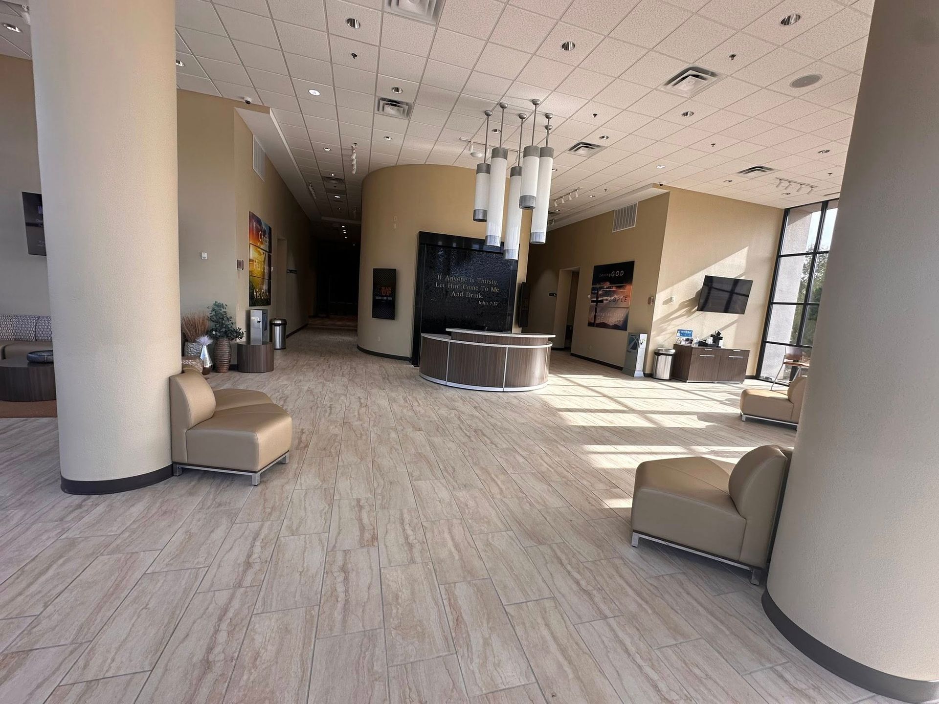 A modern, beige-toned hotel or office lobby featuring tan armchairs, a curved front desk, and a tiered light fixture.