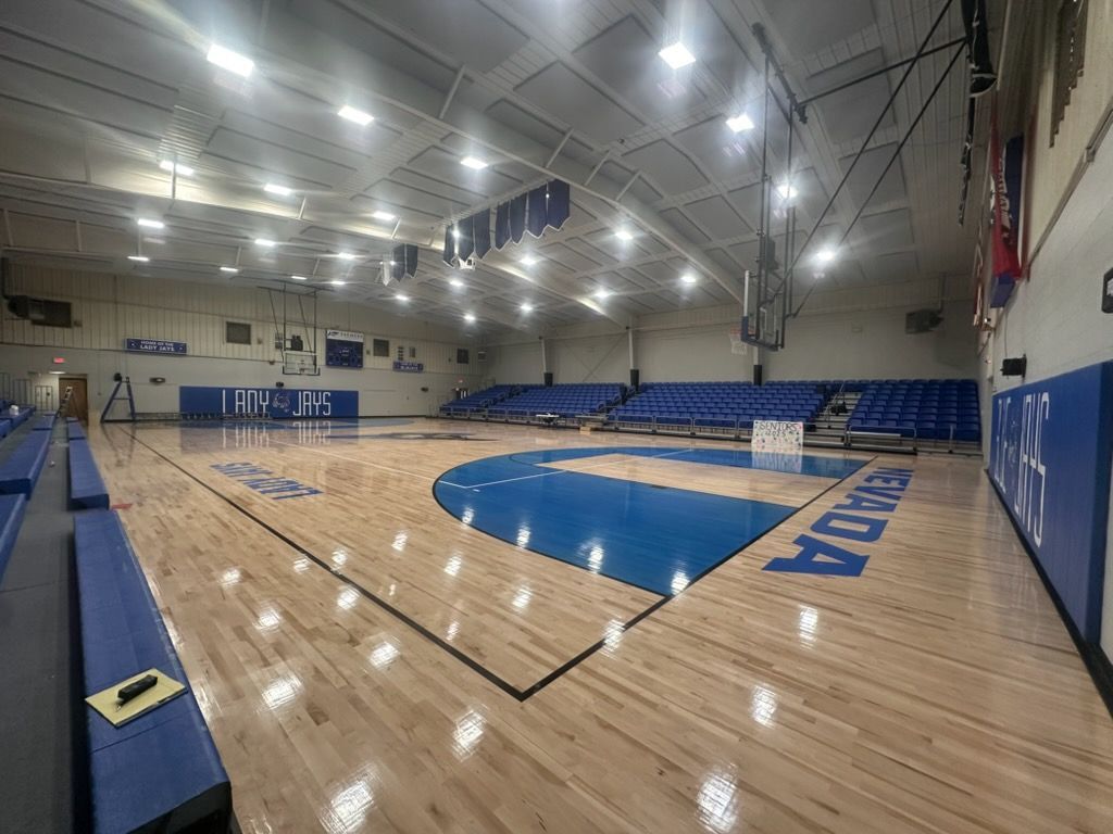 An indoor basketball court with polished wood floors, blue painted zones, and blue tiered bleachers along the wall.