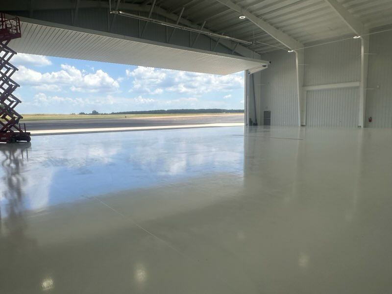 A spacious hangar with a highly reflective, glossy white floor and an open door looking out onto an airfield.