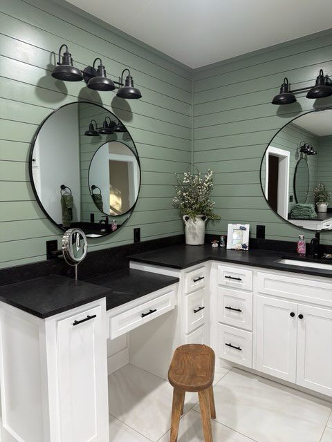 A sage green shiplap bathroom vanity with white cabinets, black countertops, two round mirrors, and matching sconces.