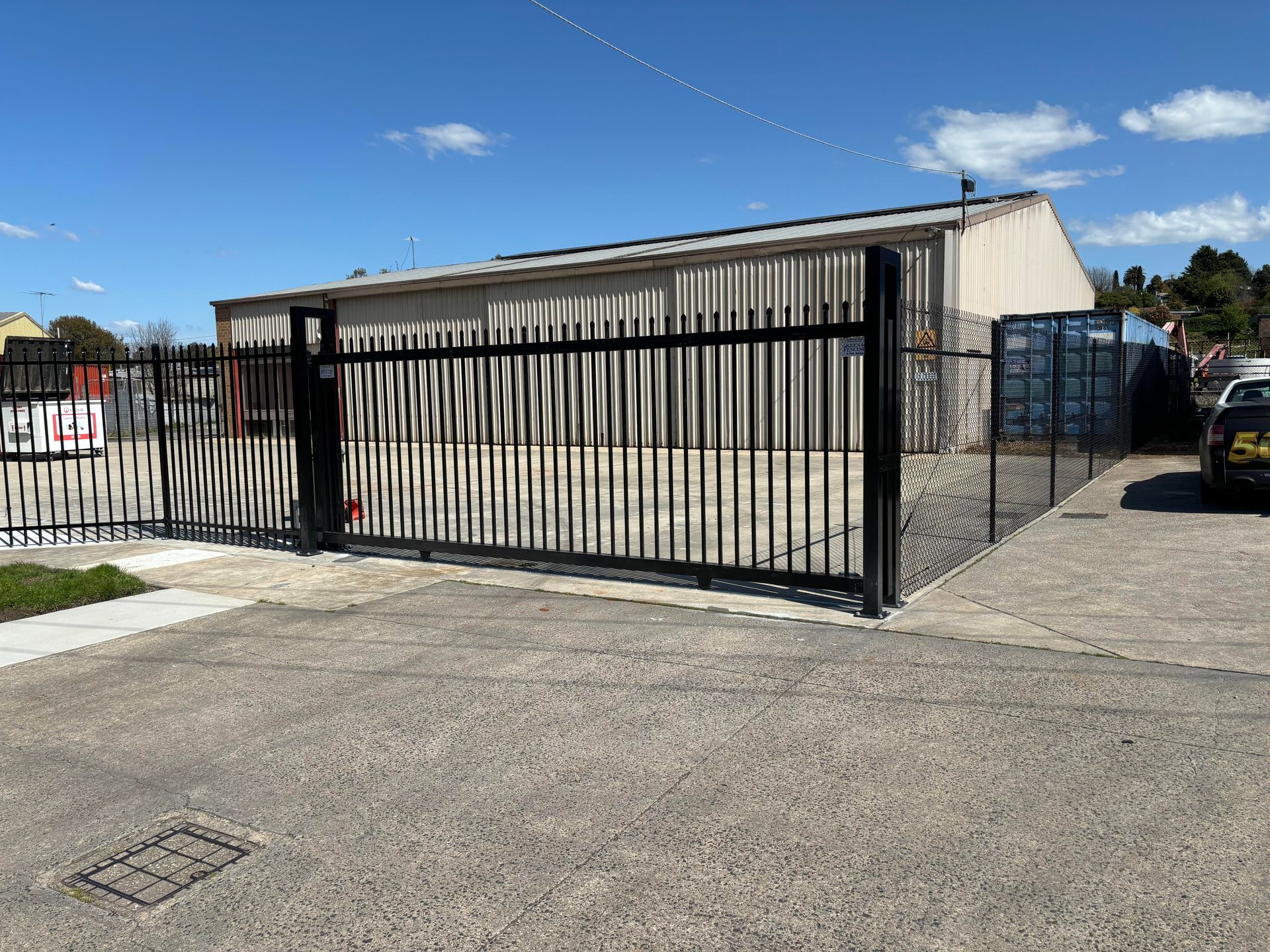 Black palisade sliding metal gates in front of a beige warehouse on a sunny day.