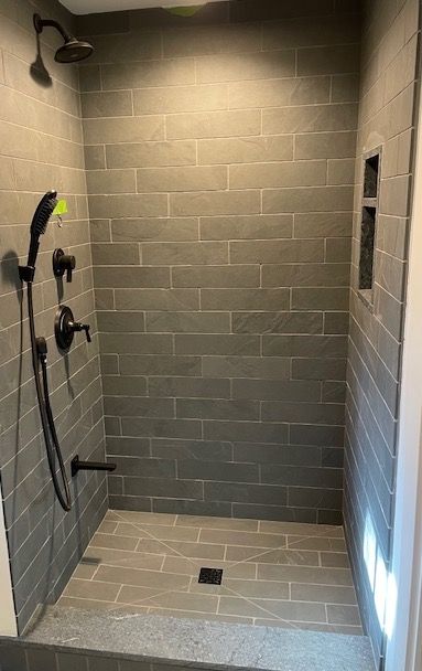 A walk in shower with gray tiles and a black shower head.