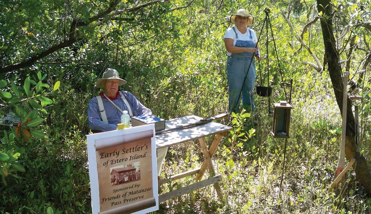 Two people at a woodland trailhead kiosk, one seated with papers and one standing beside a table.