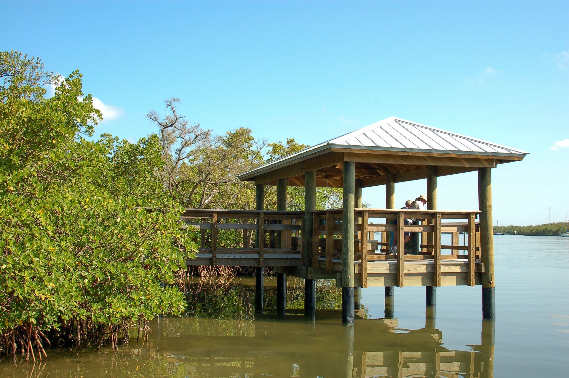 Wooden gazebo on stilts beside calm water, with green trees and a clear blue sky.