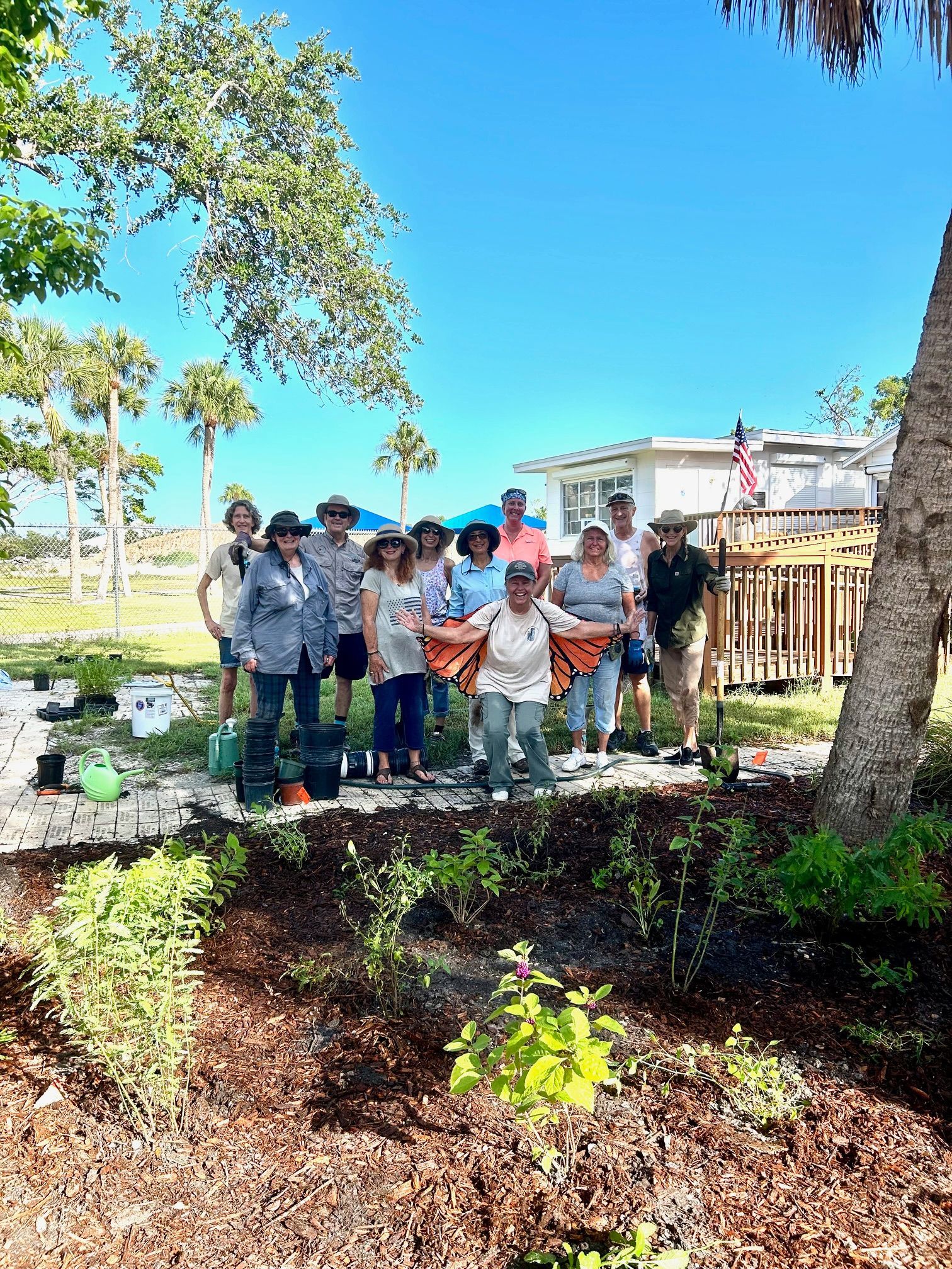 Group of people posing outdoors beside a tree and garden bed under a bright blue sky