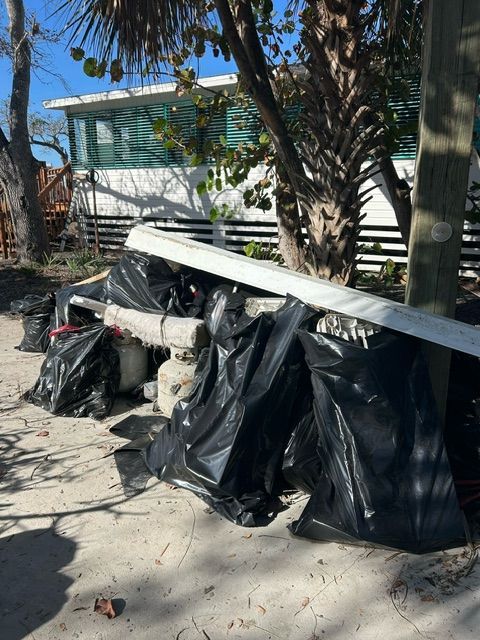 Black trash bags and white boards stacked beside a palm tree on a sunny sidewalk.