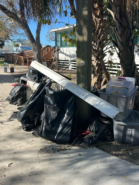 Piles of trash bags and bulky debris on a sidewalk beside a tree and fence in a sunny yard