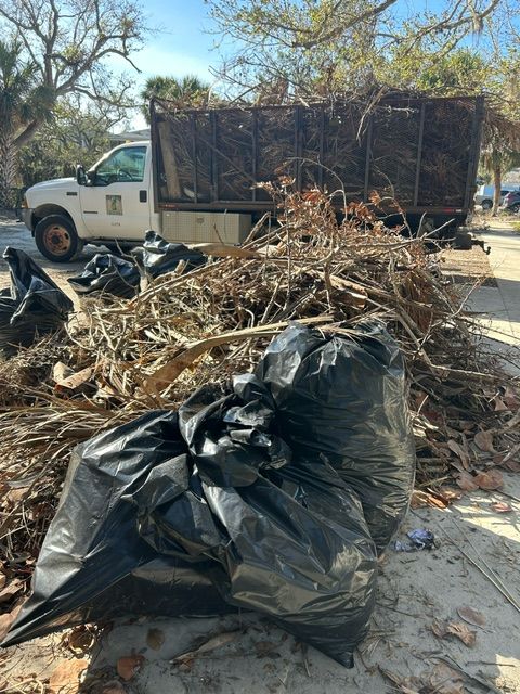 Black yard-waste bags and branches piled beside a white truck in a sunny outdoor area
