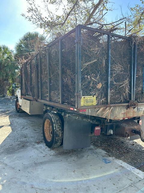 Flatbed truck with a large wire cage trailer parked along a roadside under trees.