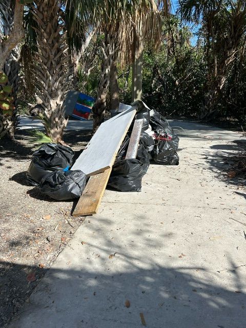 Long white plank leaning on black trash bags along a palm-lined sandy path