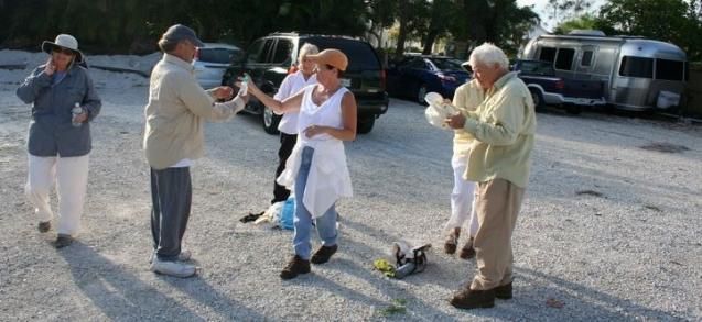 Four people outdoors on a gravel lot, clapping and talking near parked vehicles.