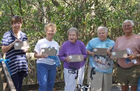 Five people standing outdoors, smiling and holding craft projects in a wooded area.