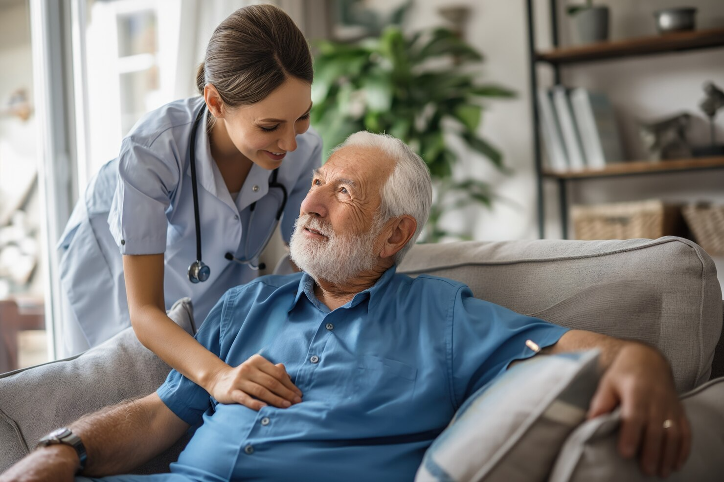 An elderly man is sitting on a couch with a nurse standing next to him.