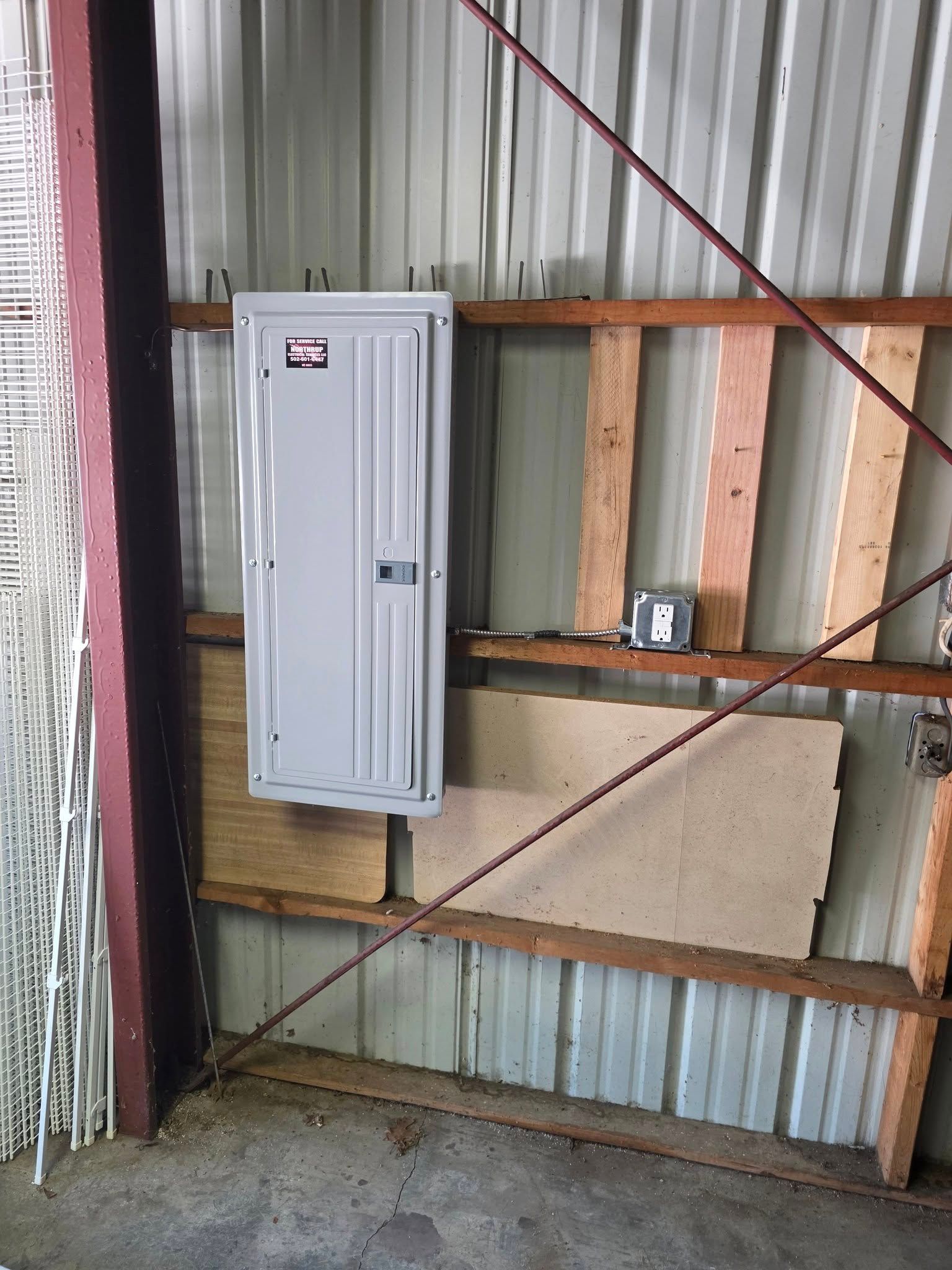 Gray electrical panel on a metal wall, supported by wooden boards. Red support beams.