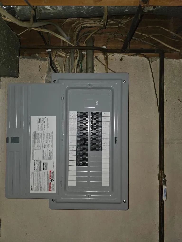 Gray electrical breaker panel mounted on a basement wall with exposed wiring overhead.