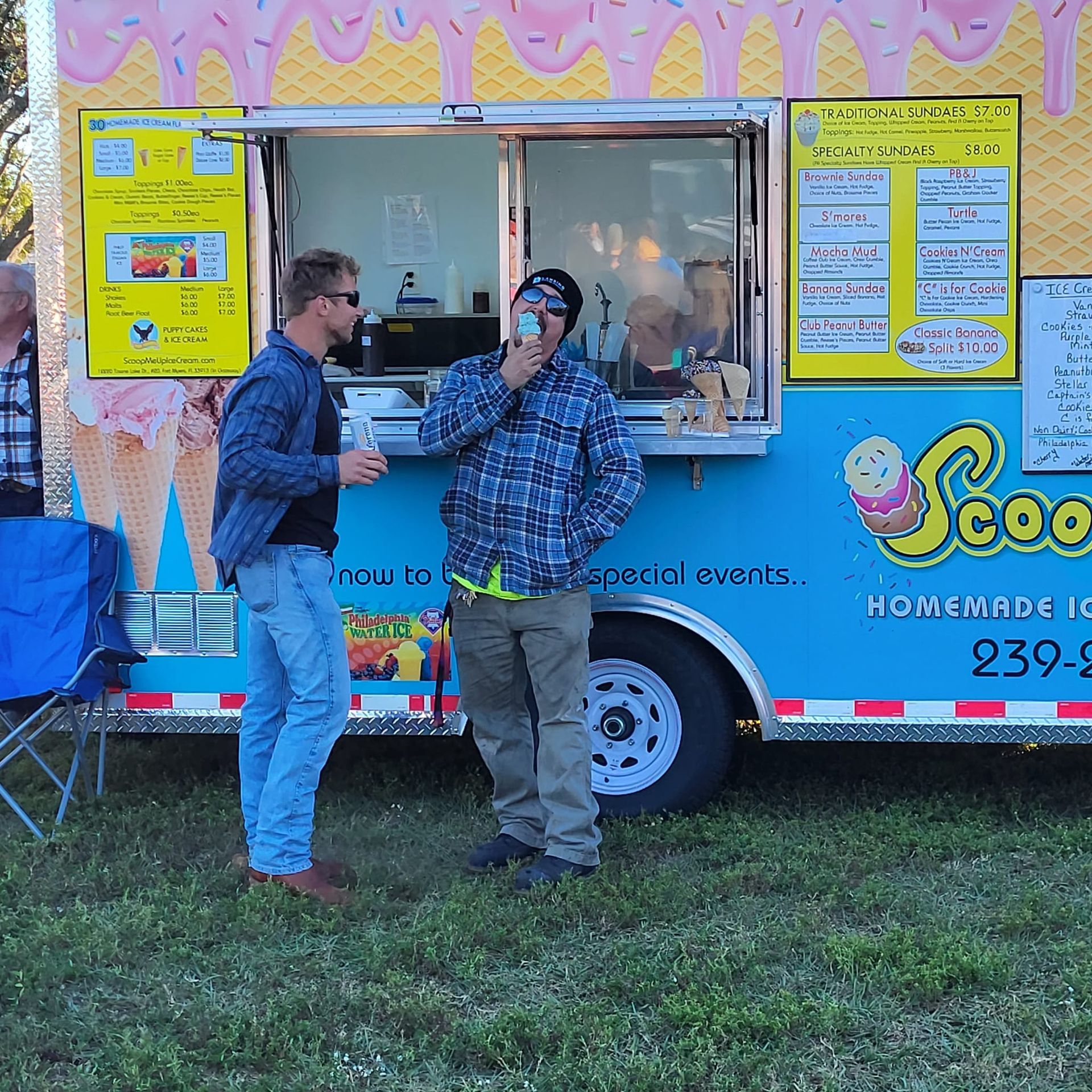 Two men stand near a blue ice cream truck, one drinking a beverage. Outdoors, grass setting.