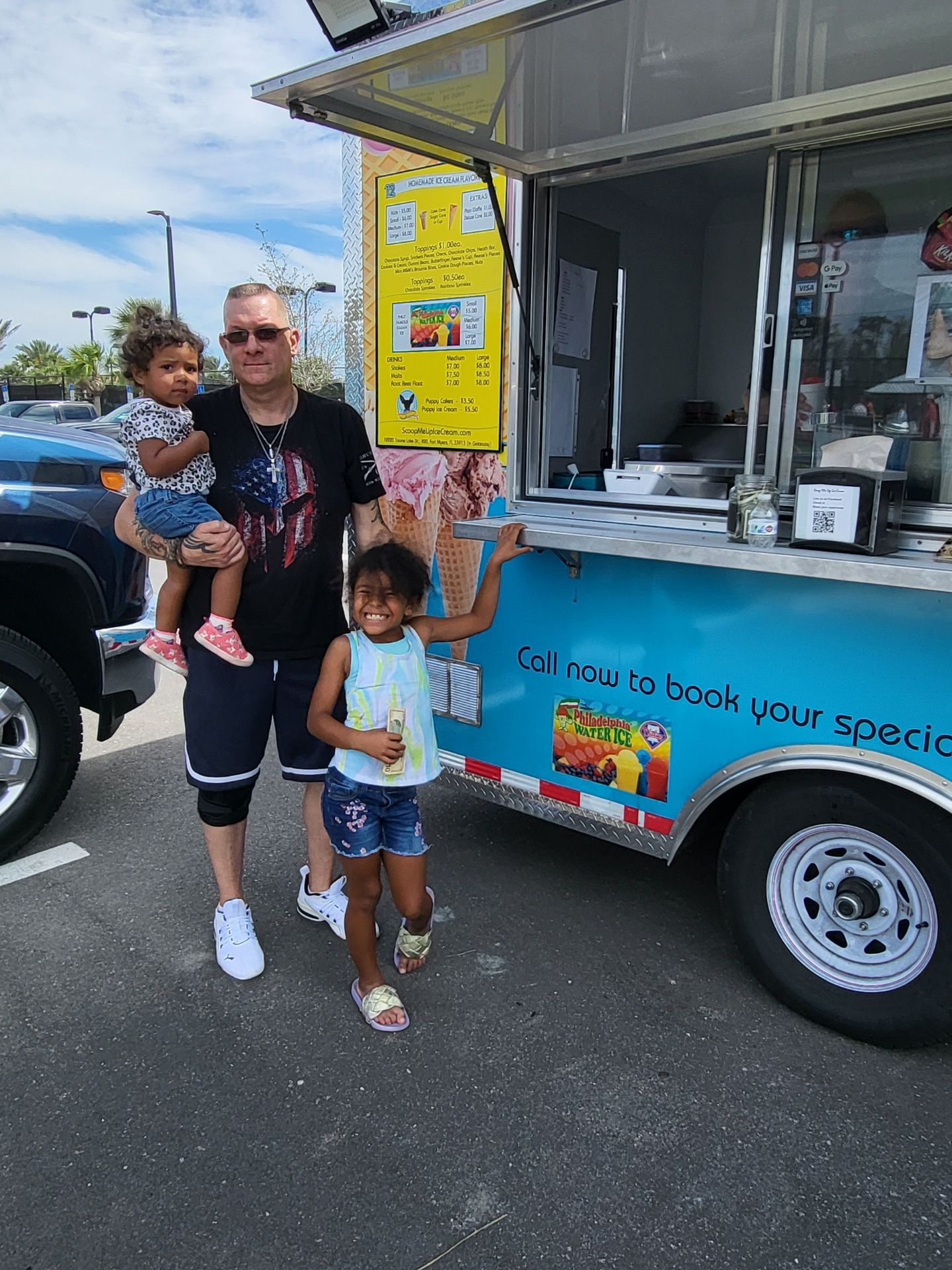 Man holding child, standing with a girl in front of a blue food truck. Sunny day.