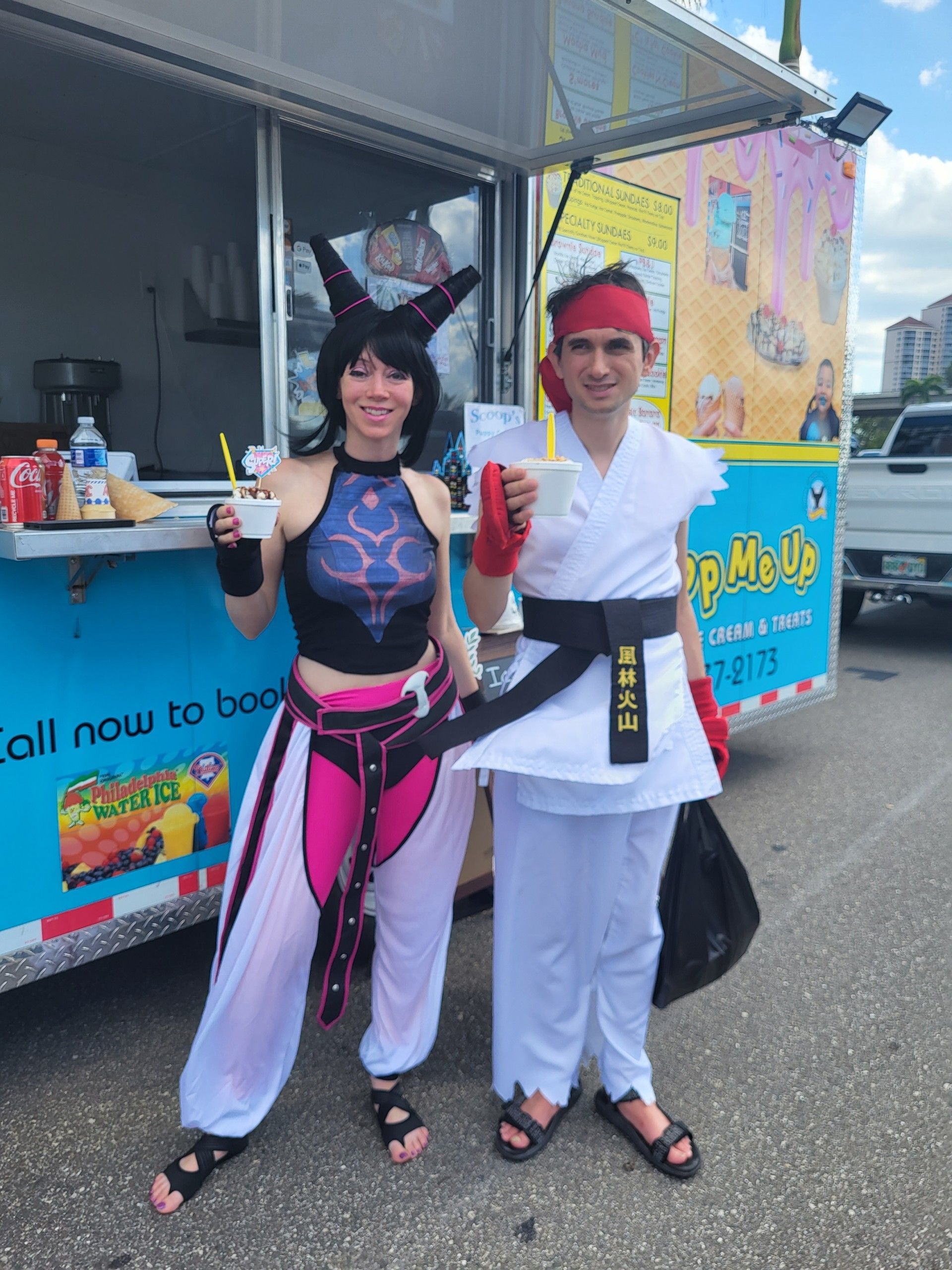 Two cosplayers in front of a food truck. One as Juri Han, the other as Ryu, each holding a drink.
